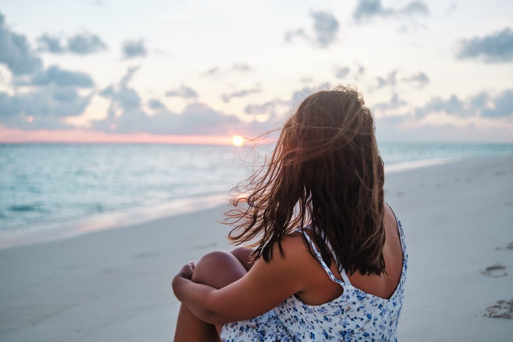 A woman sits on the sand looking toward the ocean at sunset on a beach in the, Maldives.