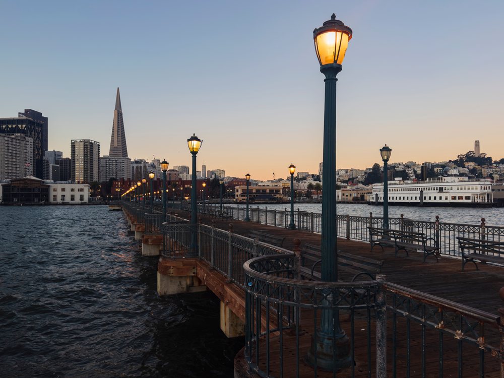 Wooden Pier with lit lamp posts and benches overlooks the water and city skyline including the Transamerica Building at dusk in Embarcadero, landscape, cityscape, city life, San Francisco, California, USA.