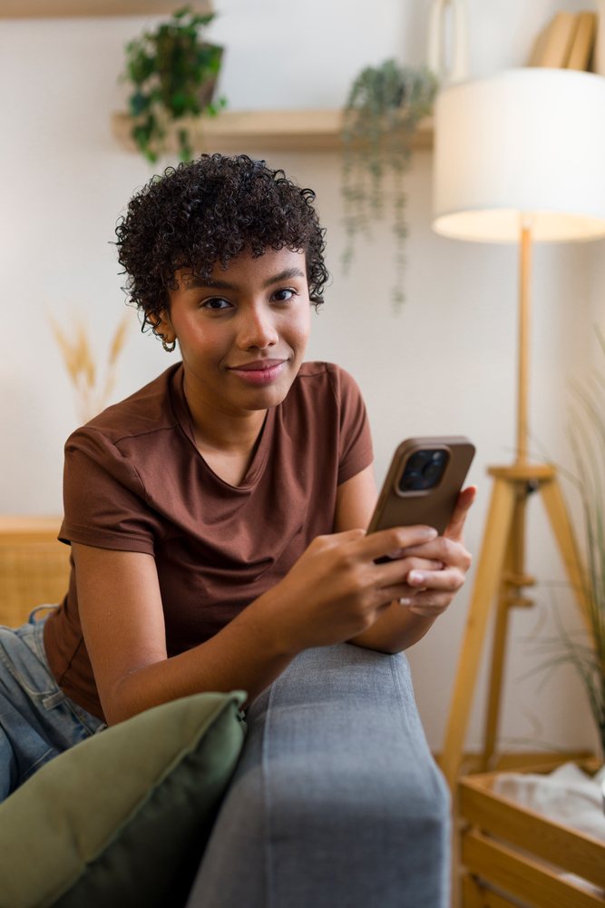 Young woman with curly hair smiling while sitting on a couch holding a smartphone at home.