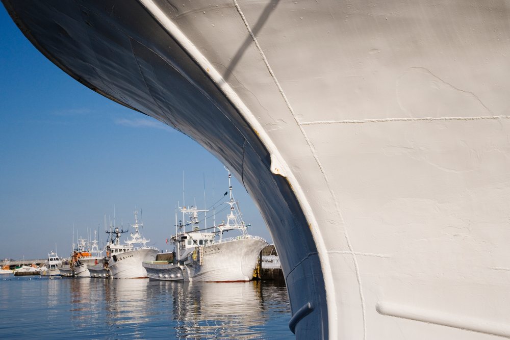 Large white ship bow in the foreground with several moored fishing boats in a calm harbor under a clear blue sky, Hokkaido, Japan.