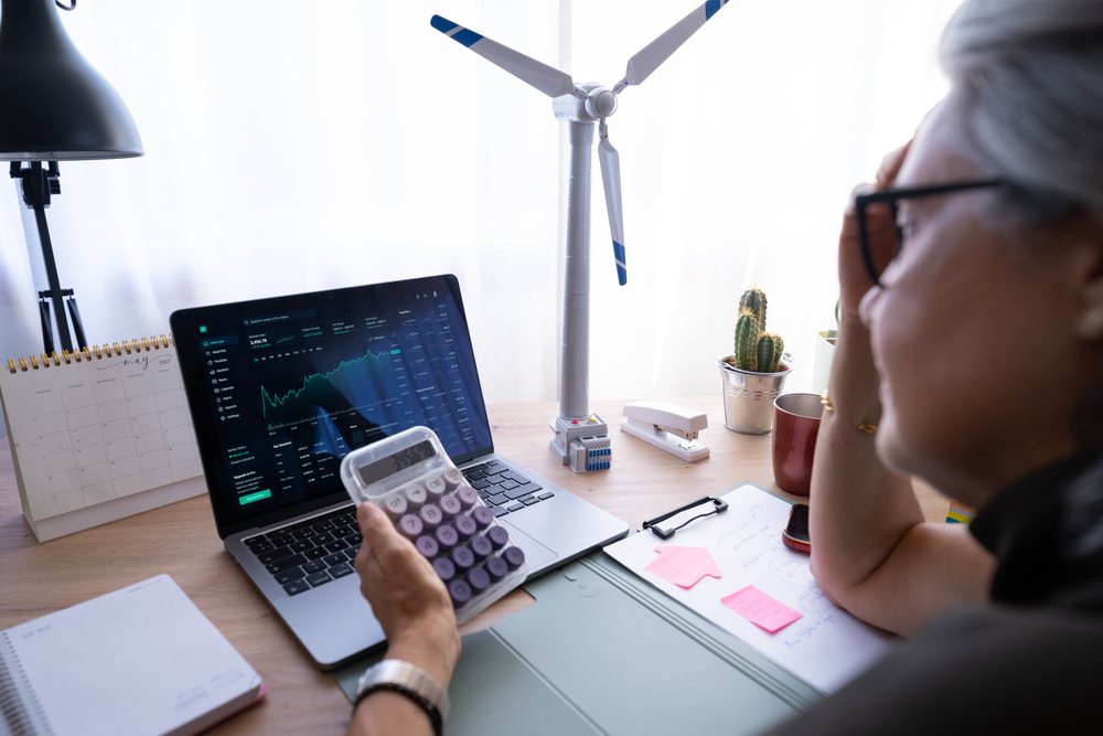 Woman working at her desk with calculator and financial data on laptop screen near wind turbine model and calendar, Spain.