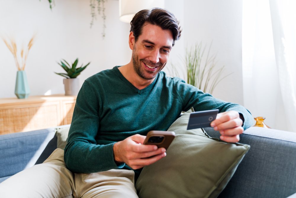 Man wearing a green sweater and beige pants is sitting on a couch holding a credit card and using his phone indoors in natural daylight, Spain.