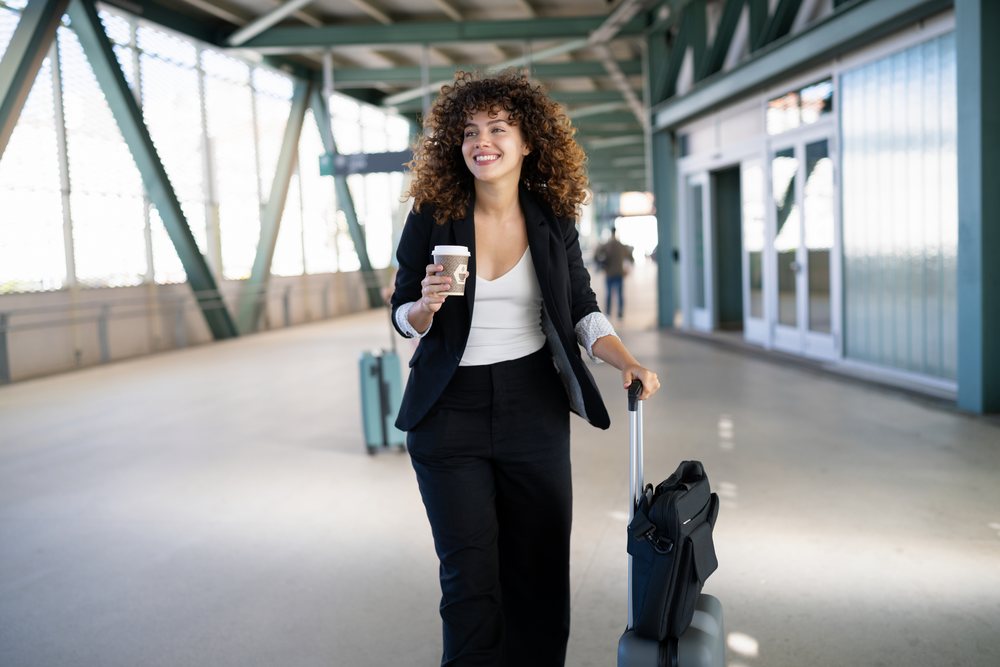 Woman in casual business attire walks through an airport terminal, pulling luggage and holding a disposable coffee cup, Spain.
