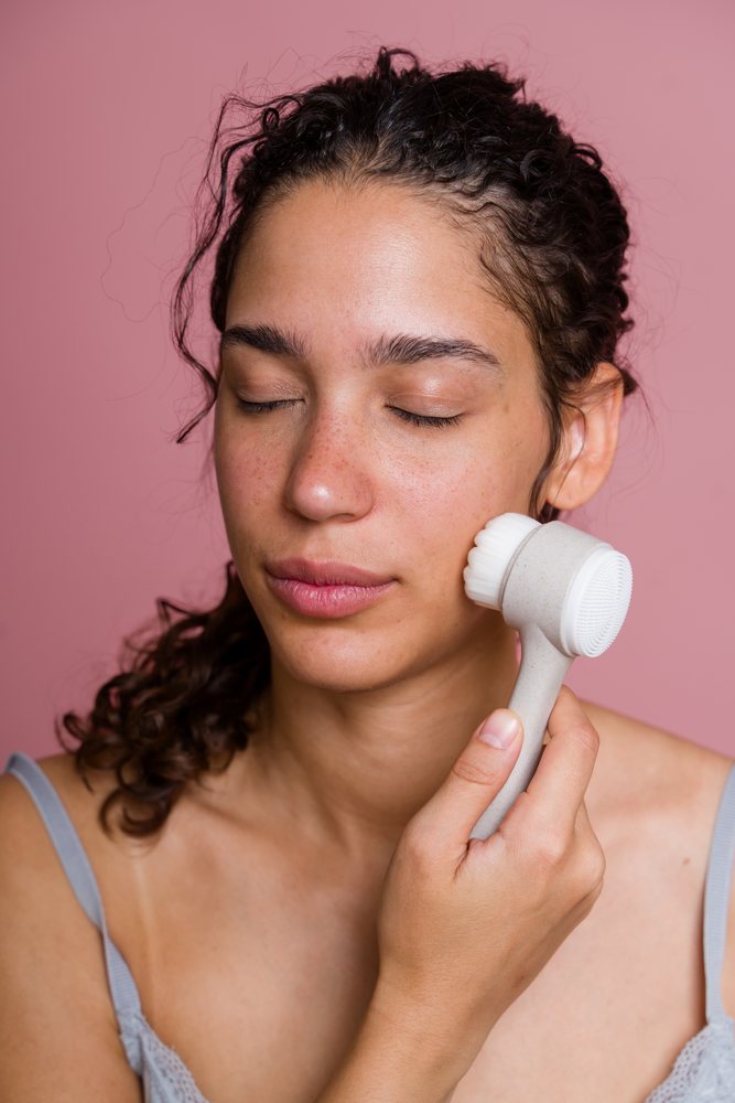 Woman with curly hair using a facial cleansing brush on her cheek against a pink background, beauty shoot, Spain.