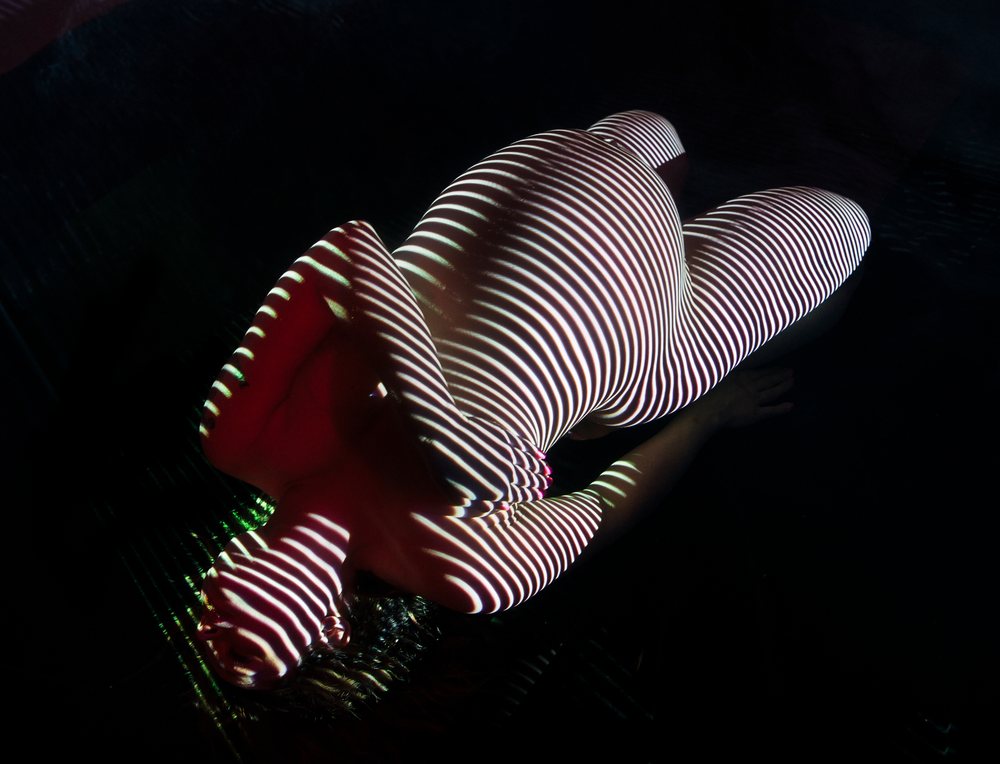 Pregnant woman lying down with striped light projections emphasizing her body curves against a dark background, pregnancy.