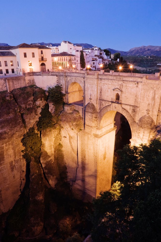 The Puente Nuevo stone bridge spans a deep gorge, with city lights and mountains visible at evening, Andalucia, Spain.