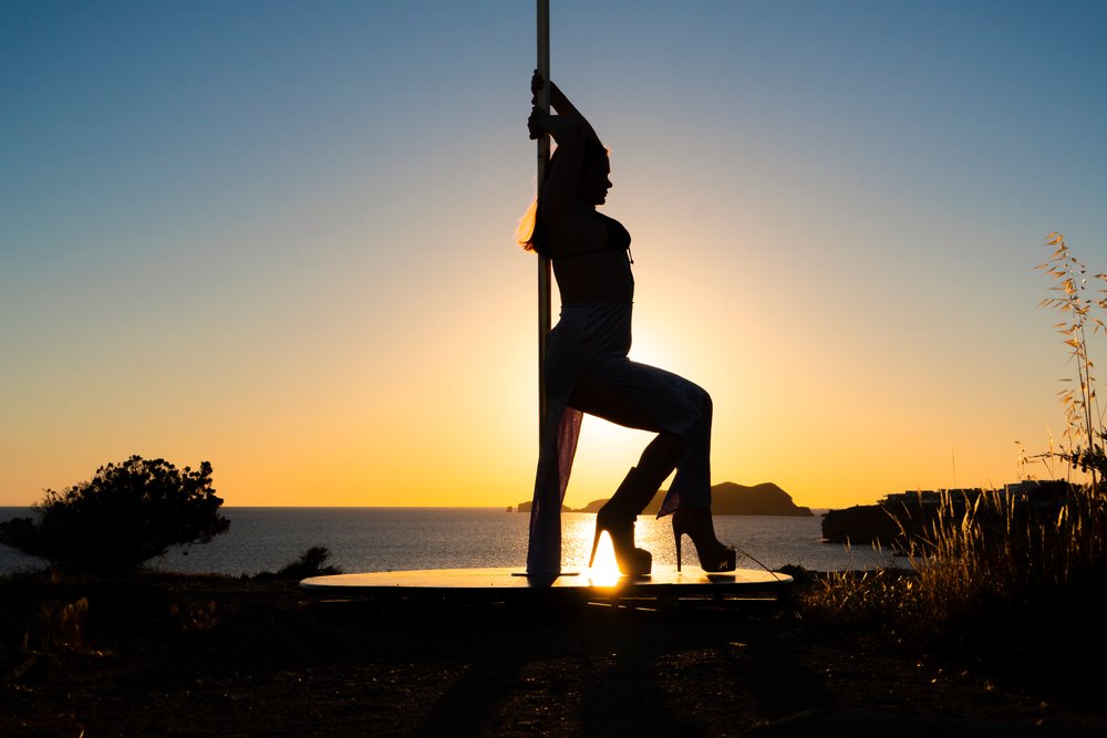 Silhouette of a woman performing pole dance on a platform at sunset with the sea and islands, Ibiza, Spain.