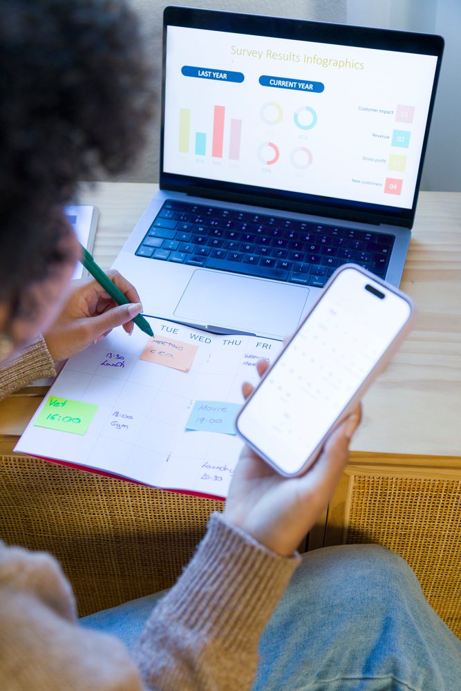 Woman sitting at a wooden desk in a home office plans her schedule using a calendar, smartphone, and laptop displaying survey infographics, remote work.