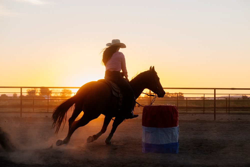 Cowgirl riding a horse around a barrel in an outdoor arena at sunset, Southwest USA.