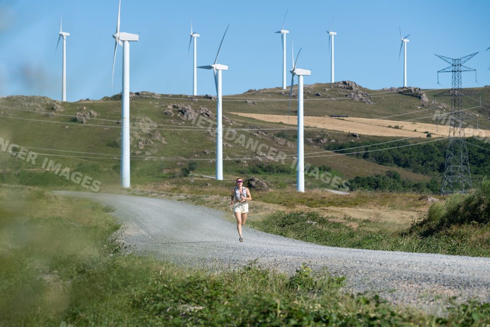 A white woman running on a gravel trail near wind turbines in Galicia