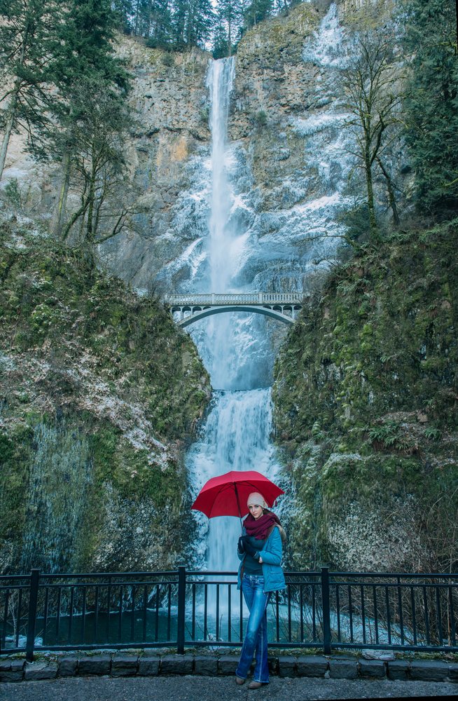 Woman in winter clothing holds a red umbrella near the two-tiered Multnomah Falls with its arched pedestrian bridge, Bridal Veil, Oregon.