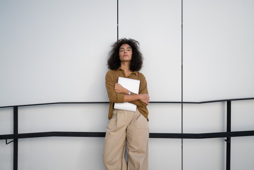 Woman standing outdoors holding a closed laptop with eyes closed against a white wall with black railing, Madrid, Spain.