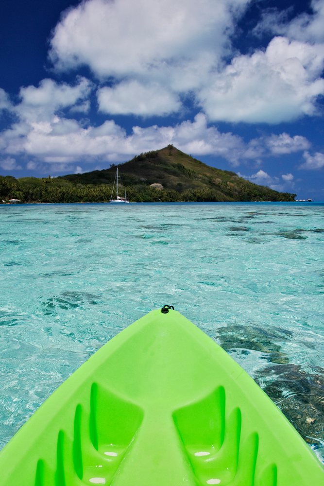 Green kayak floats in crystal clear turquoise lagoon waters facing a tropical island hill, French Polynesia under a partly cloudy blue sky, Tahiti.