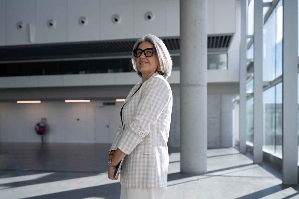 Businesswoman stands smiling in a modern office building, holding a tablet and wearing glasses and a checkered blazer, Spain, Madrid.