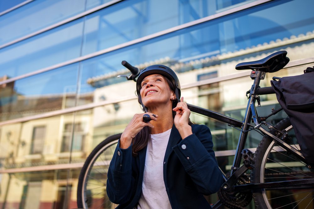 Woman adjusts her helmet strap beside a black bicycle reflecting in glass in Málaga urban setting.