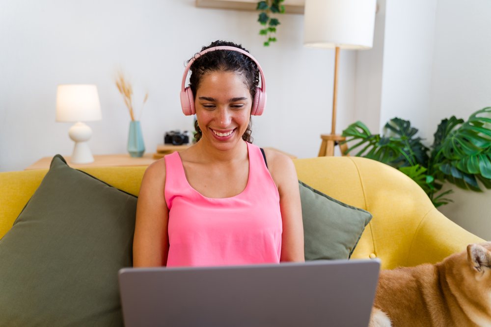 A woman wearing pink headphones and a pink tank top smiles while using a laptop on a yellow couch with a dog nearby in a living room, Spain.