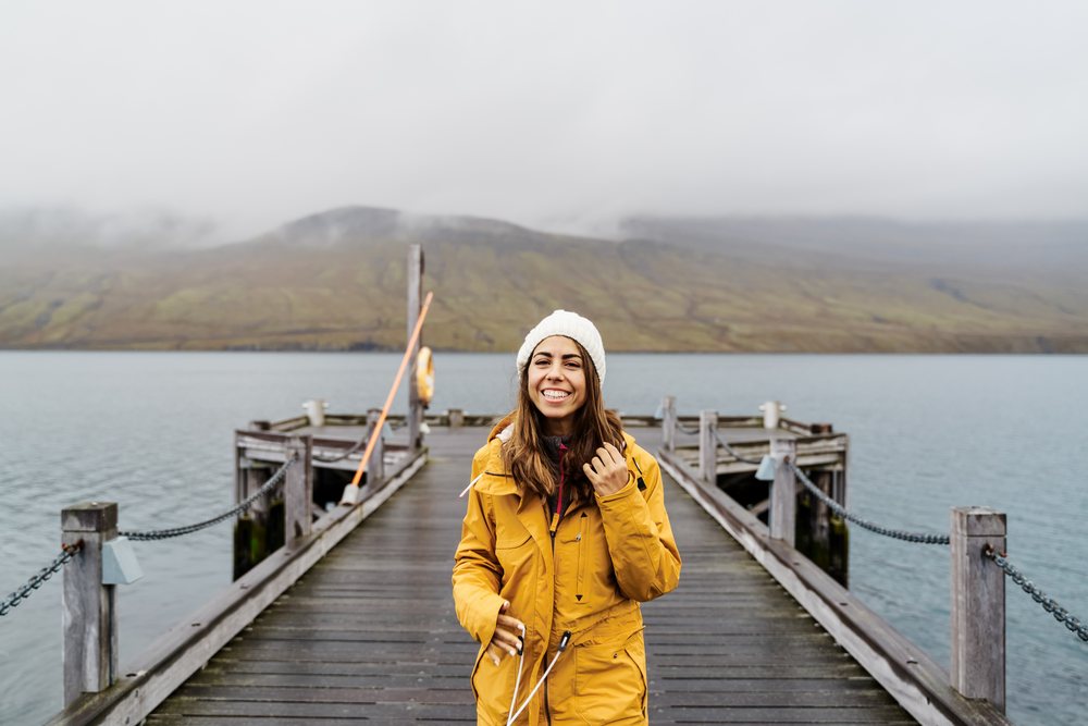 A smiling woman wearing a yellow jacket and white knit hat stands on a wooden pier by the water with misty mountains in the background, Iceland.