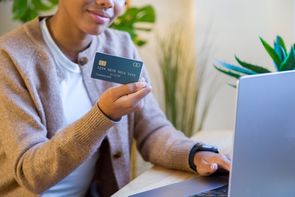 Woman holding credit card and using a laptop in a home office with a plant visible in the background, remote work.