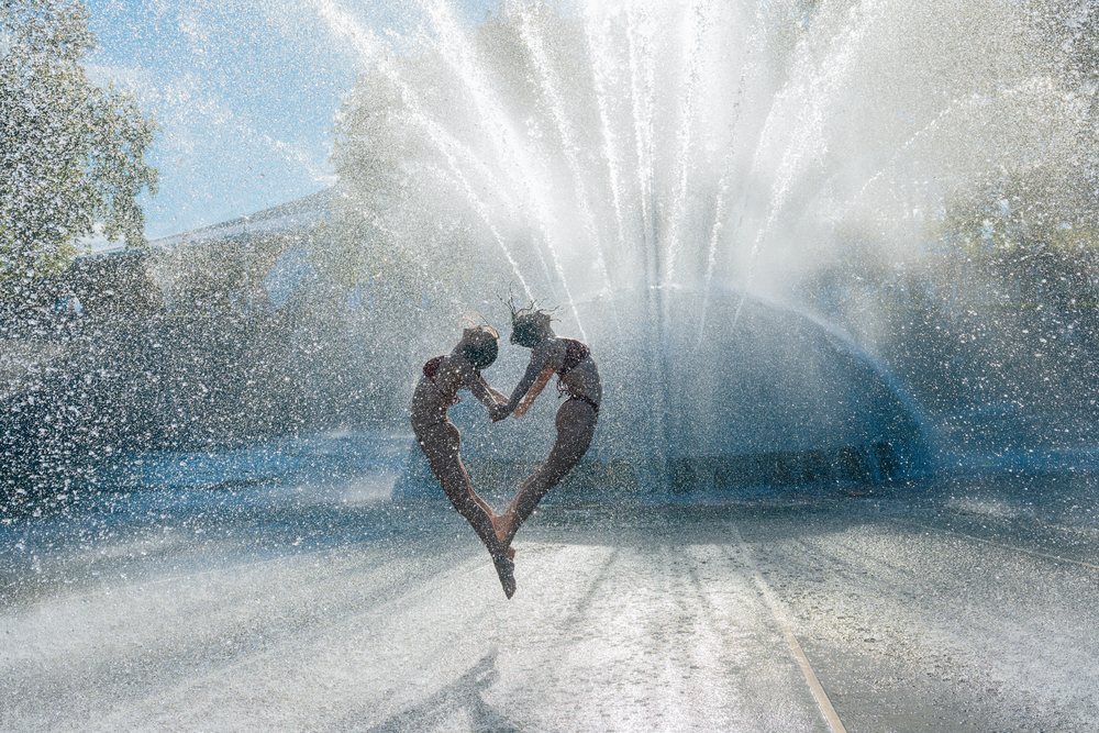 Two women in bikinis jumping and holding hands in front of a water fountain splash on a sunny summer day outdoors, partner, acrobatics, yoga, USA.