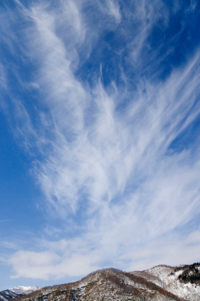 Cirrus clouds drift above snowy mountain peaks under a clear blue sky, Hokkaido, Japan.