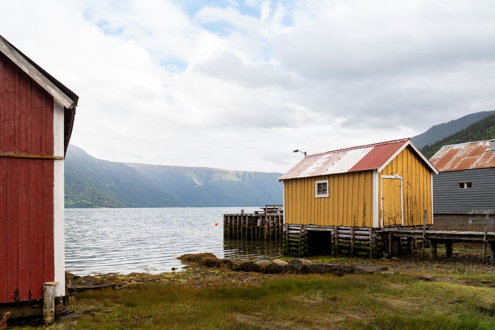 Wooden boathouses on stilts by the calm Fjord shore, with mountains and cloudy sky in the background, coastal town, village, Luster, Norway.
