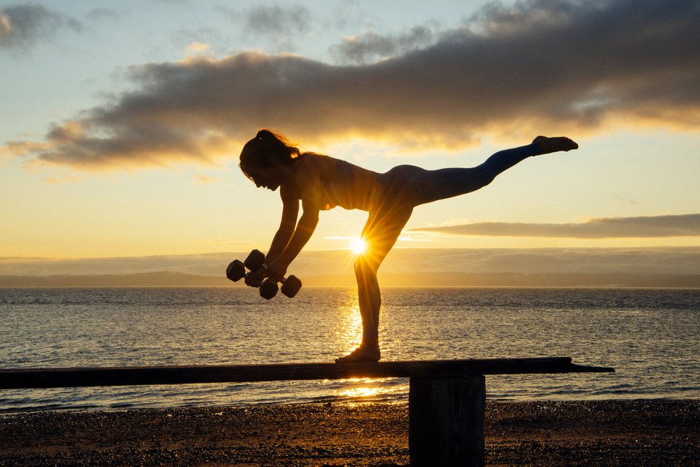 Woman balances on one leg holding dumbbells at sunset on a calm beach, WA, USA.