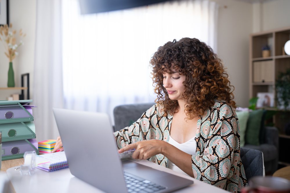 Woman with curly hair works on a laptop at her desk in a well-organized home office with stationery and plants, Spain.