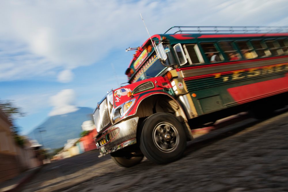 Colorful chicken bus carrying seated passengers travels downhill on a cobblestone street, with a mountainous landscape visible, Sacatepequez Department, Guatemala.