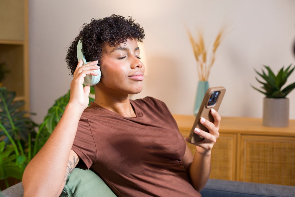 Young woman with curly hair wearing headphones and holding a smartphone seated indoors at home.