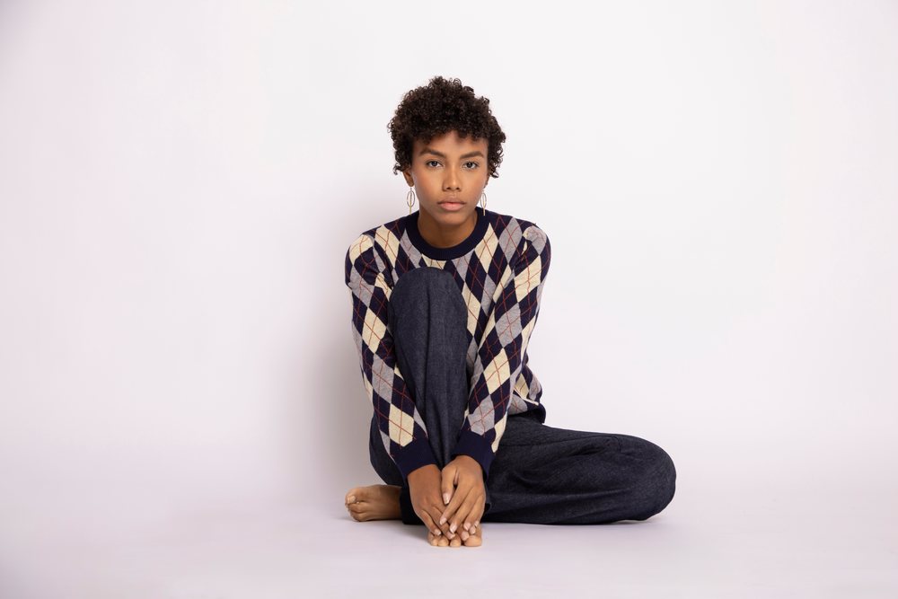 Young woman sitting barefoot on the floor against a white studio background wearing an argyle sweater and dark pants, fashion, young culture.