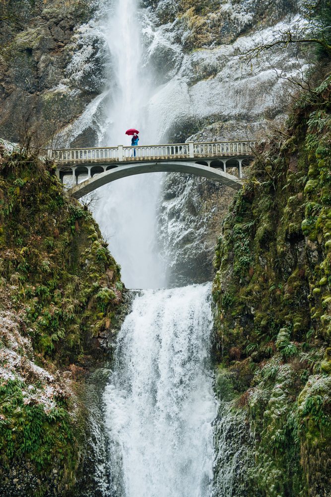 Person holding a red umbrella stands on a bridge above Multnomah Falls waterfall, Bridal Veil, Oregon.