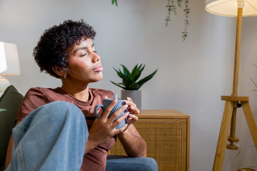 Woman seated indoors holding a ceramic coffee cup with spoon in a cozy living room with lamp and houseplant, Young woman, home.