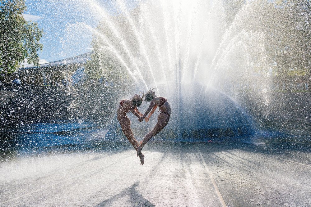 Two individuals performing a synchronized acrobatic yoga jump with hands held in front of a water fountain spray on a sunny day, partner, acrobatics, USA.