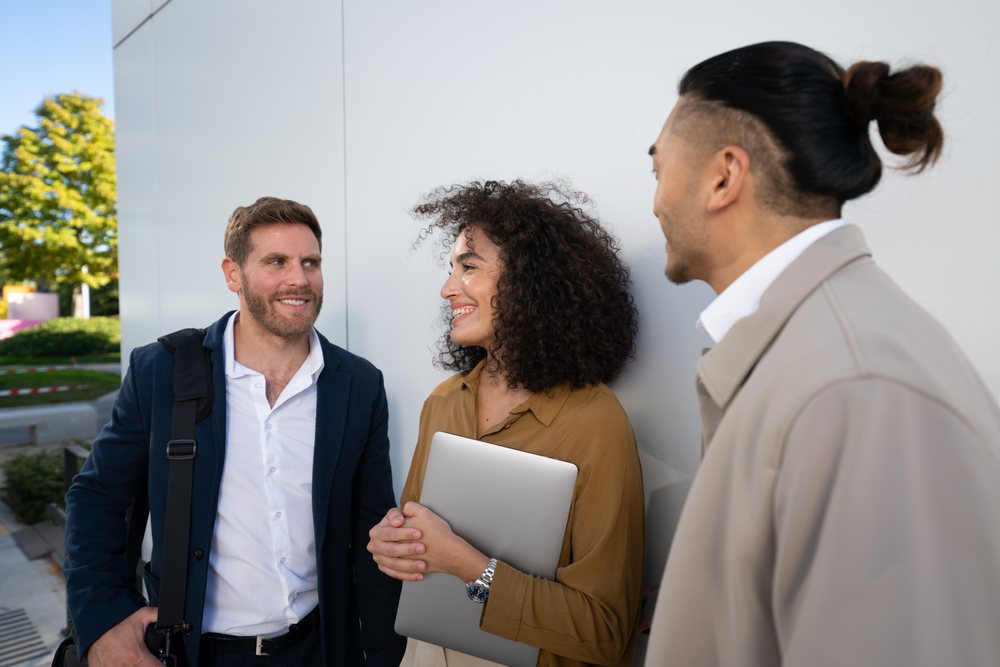 Three people in business casual attire talk and smile together outdoors near a white wall on a sunny day, Madrid, Spain.