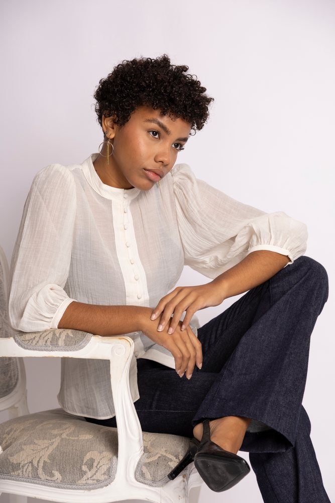 Woman with curly hair sitting thoughtfully on a white chair in a studio wearing a long sleeve white blouse and dark pants, fashion, young culture.