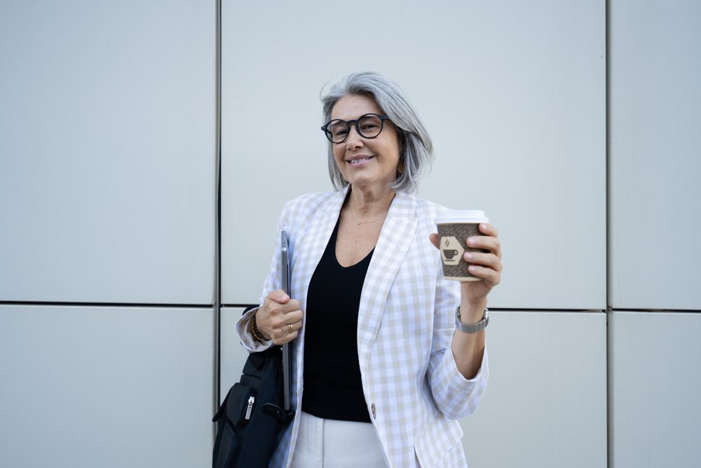 Smiling middle-aged woman with gray hair holding a coffee cup and laptop outdoors, businesswomen, Spain.