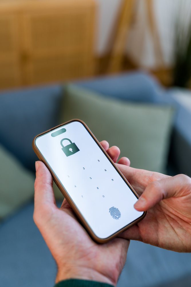 Hand holding a smartphone showing a lock screen with numeric keypad and fingerprint icon indoors with couch and pillows, Spain.