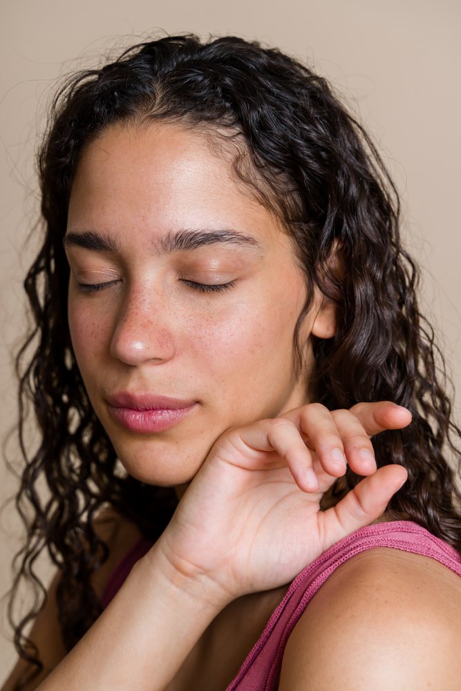 Young woman with closed eyes and curly hair posing with hand near face wearing a pink sleeveless top in a studio portrait, beauty shoot, Spain.