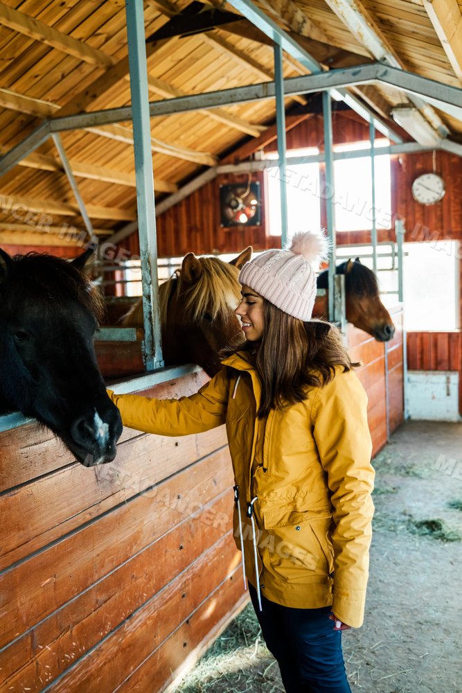A white woman pets horses inside a wooden stable barn