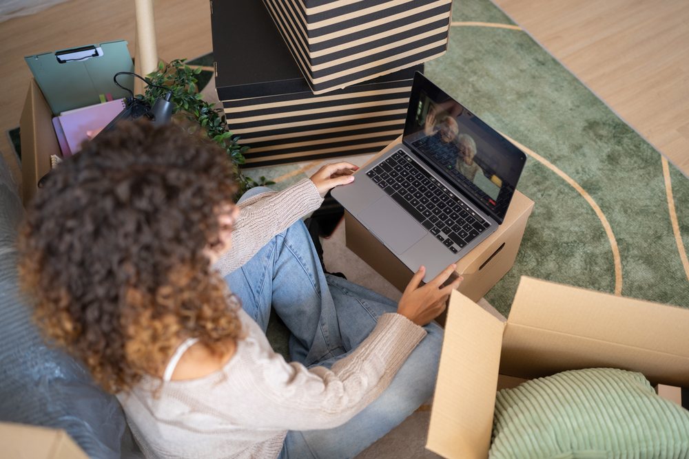 Woman sitting on green striped carpet among cardboard boxes holding a laptop showing a video chat indoors during moving, Spain.