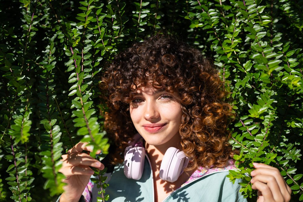 Smiling woman with curly hair wearing headphones outdoors among green foliage in natural sunlight, Spain.