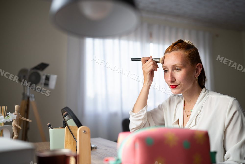 A white woman applies makeup at a vanity in a cozy home setting