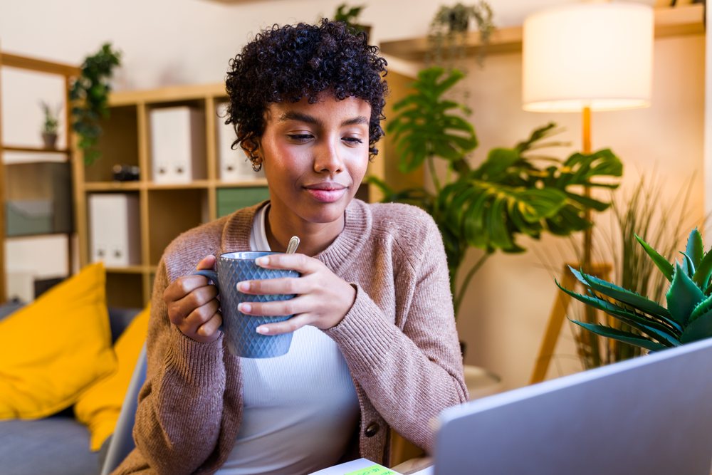 Woman holding a mug and drinking coffee indoors near a laptop and plants in a cozy home setting, Spain.