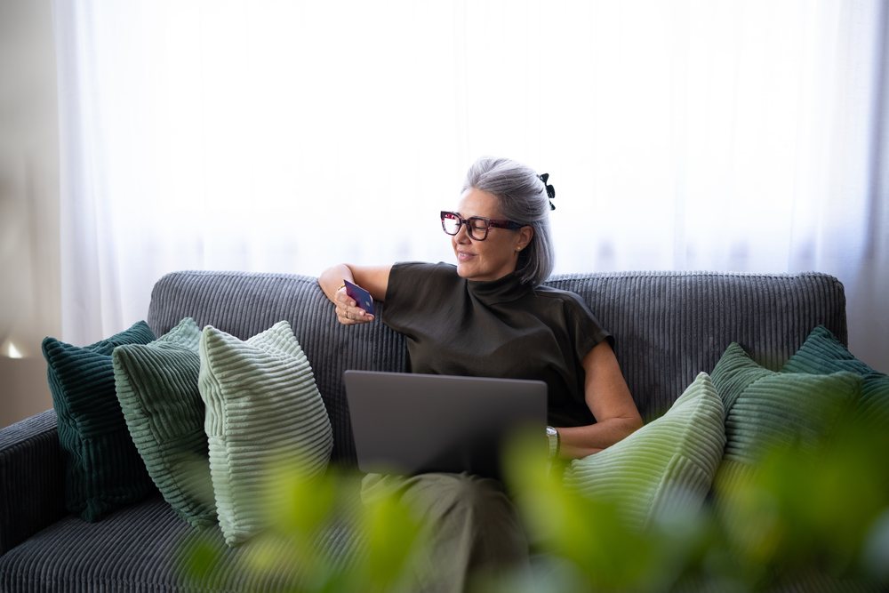 Mature woman with gray hair and glasses sits on a sofa indoors using a laptop and holding a credit card, Spain.