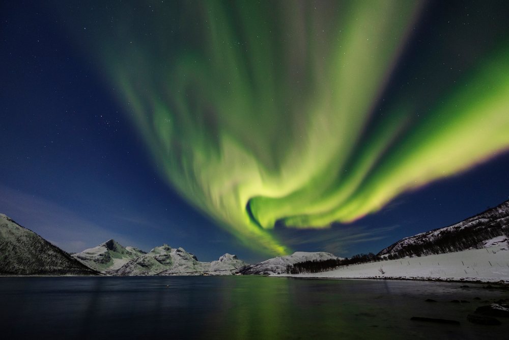 Green Northern Lights glow over snow-covered mountains and calm water reflecting the aurora, Landscape, Kvaløya, Norway.