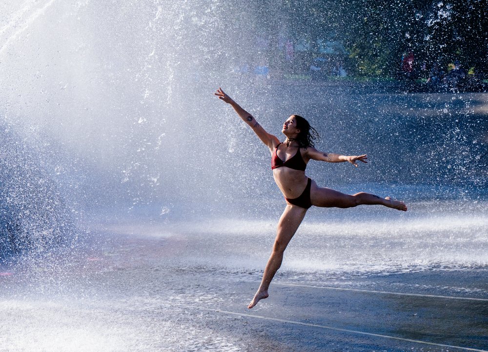 Woman jumping gracefully with arms extended amidst water splashing from a fountain in an outdoor setting, partner, acrobatics, yoga, USA.