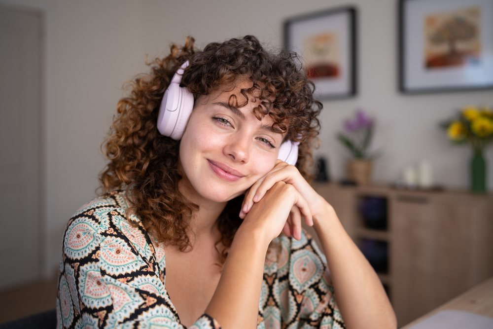 Smiling woman with curly hair wearing headphones sits indoors in a decorated home environment, Spain.
