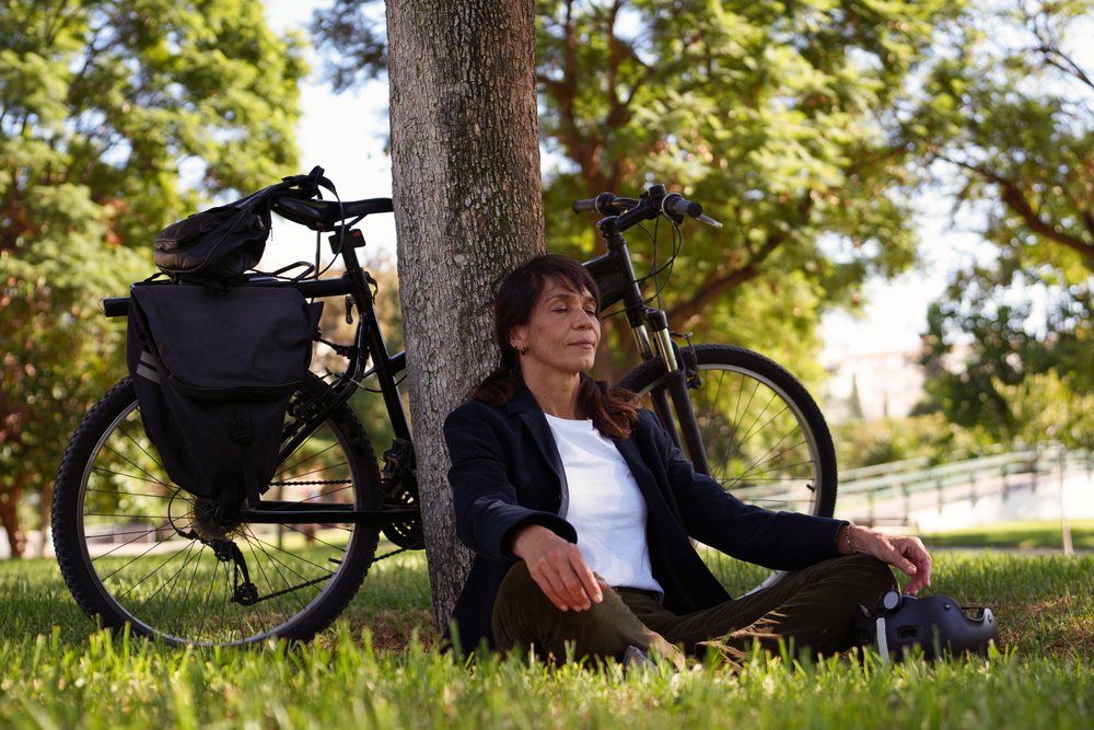 Woman meditates sitting cross-legged against a tree in a Málaga park beside a black bicycle with a helmet nearby.
