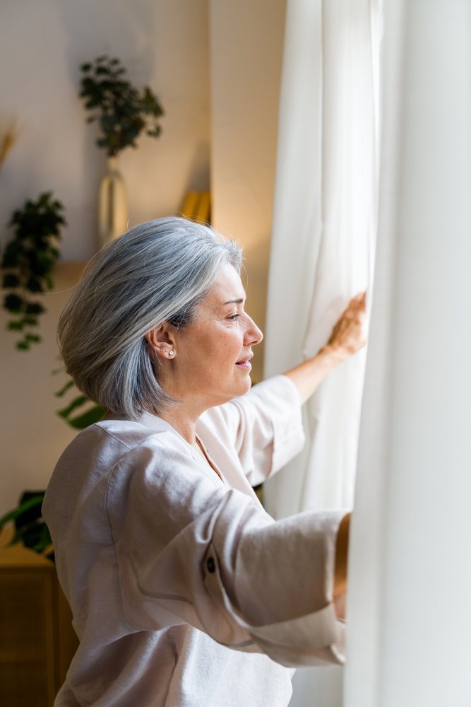 Middle-aged woman with gray hair opening white curtains to natural daylight in a cozy living room interior decorated with plants and vases, Spain.