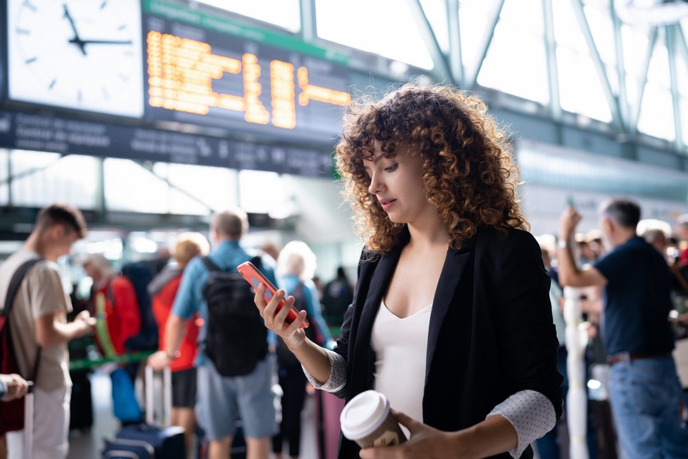 Woman holding coffee cup and checking smartphone while waiting queue near boarding display, Spain.